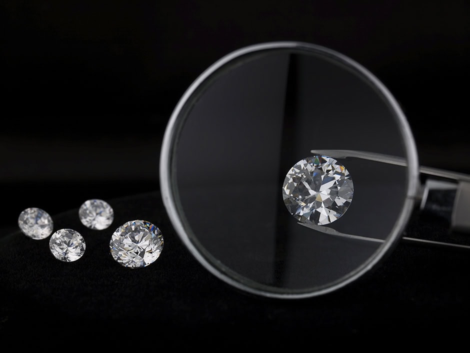 Diamond authentication: A close-up of four round, sparkling diamonds on a dark surface. A magnifying glass is focused on one diamond held by tweezers, highlighting its facets and clarity. The background is black, emphasising the diamonds' brilliance.