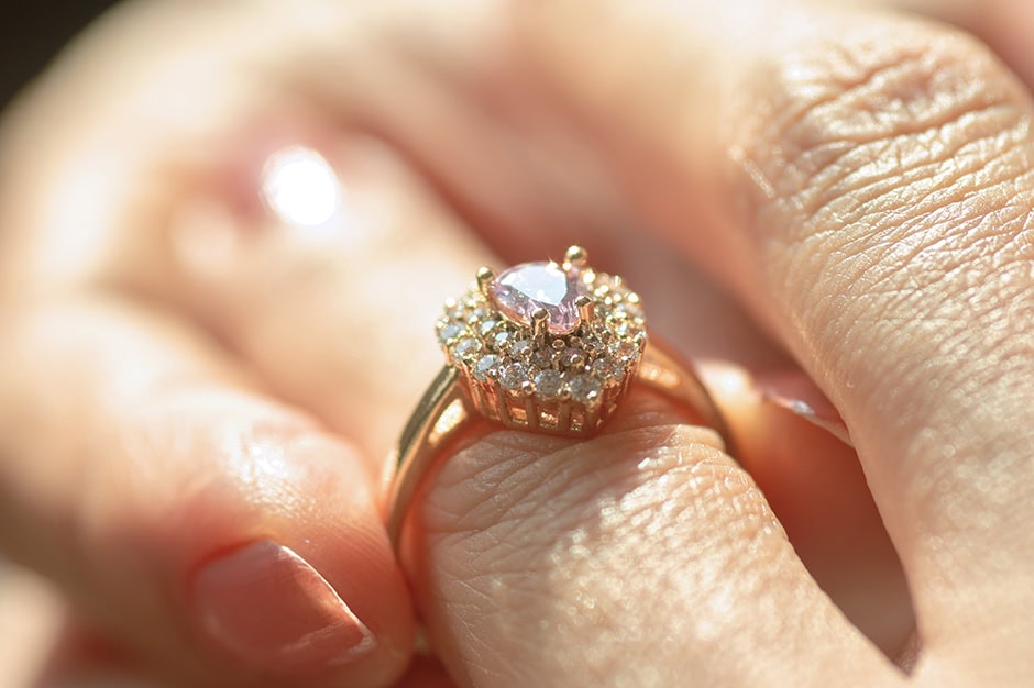 A family heirloom: A close-up of a hand wearing a gold ring, featuring a pink gemstone surrounded by small diamonds. The hand has a light skin tone and well-manicured nails. The ring sparkles in the light.