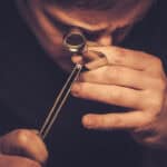 A person closely examines a small gemstone with a jeweller’s loupe, holding the gem with tweezers and using focused light to inspect its details.