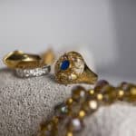 Jewellery suitable for online valuation: A close-up of gold jewelry, including a gold ring with a blue stone and an ornate design, a gold band with embedded gems, and a beaded necklace, all displayed on a textured beige surface.
