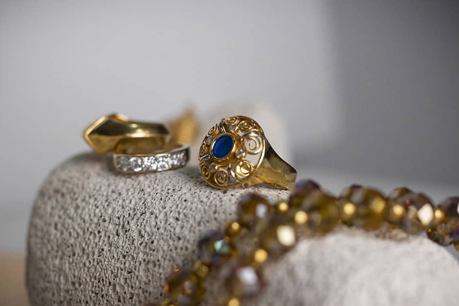 Jewellery suitable for online valuation: A close-up of gold jewelry, including a gold ring with a blue stone and an ornate design, a gold band with embedded gems, and a beaded necklace, all displayed on a textured beige surface.
